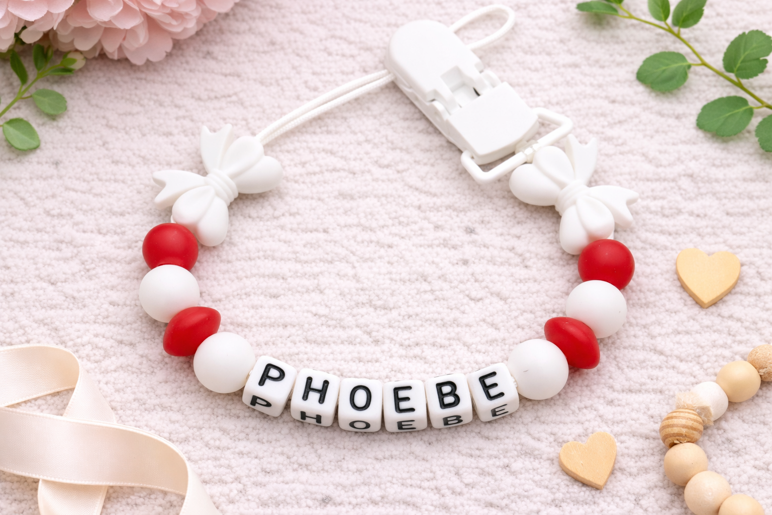 Flat lay image of a personalised baby pacifier clip featuring the name “PHOEBE” in white letter beads. The design includes white butterfly-shaped silicone beads, red and white round beads, and a white clip, styled on a soft neutral textured background with decorative flowers, greenery, ribbon, and small wooden heart accents.