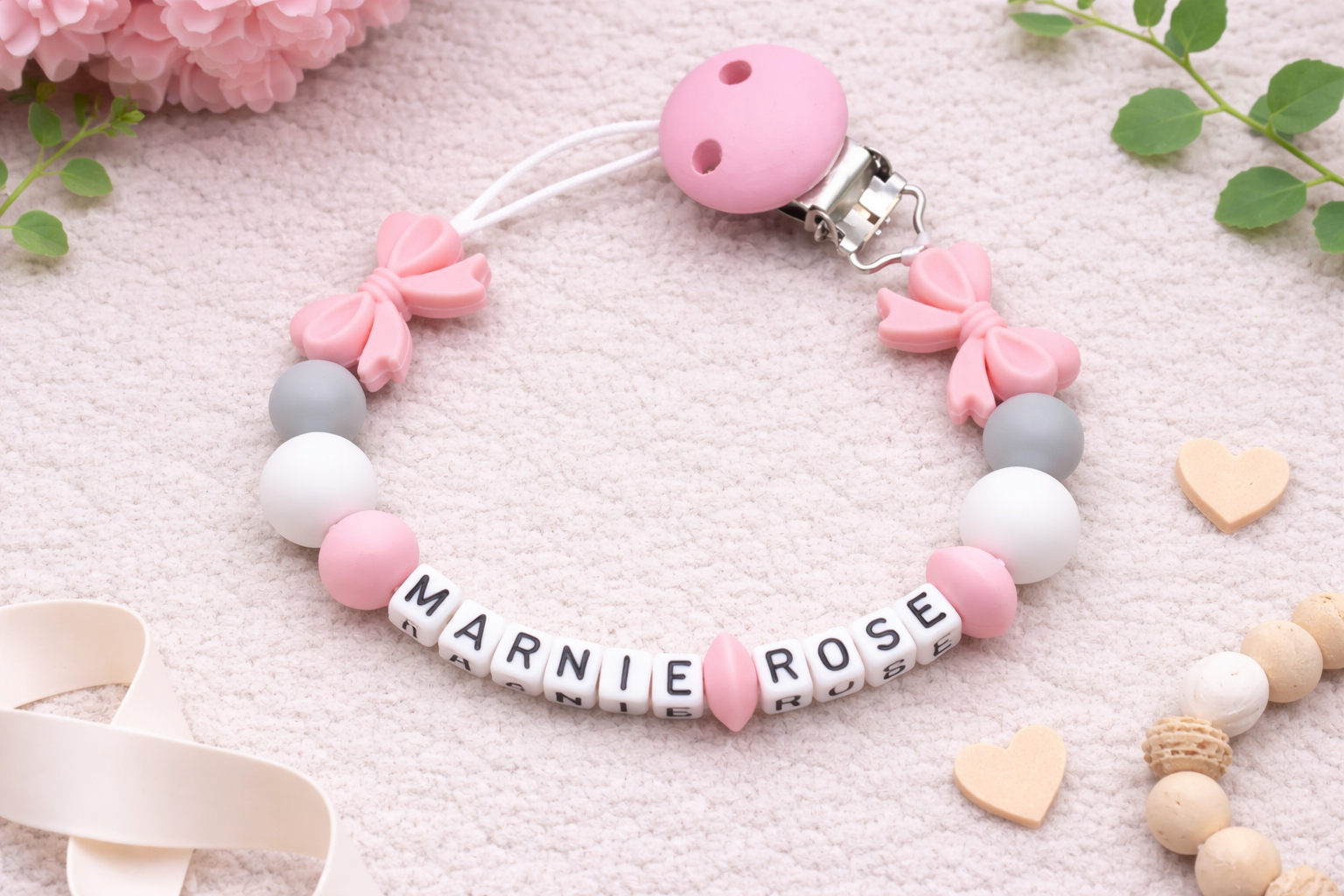 Personalised pink and grey pacifier clip with bow beads and the name Marnie Rose displayed on a soft cream flat lay background with floral accents.