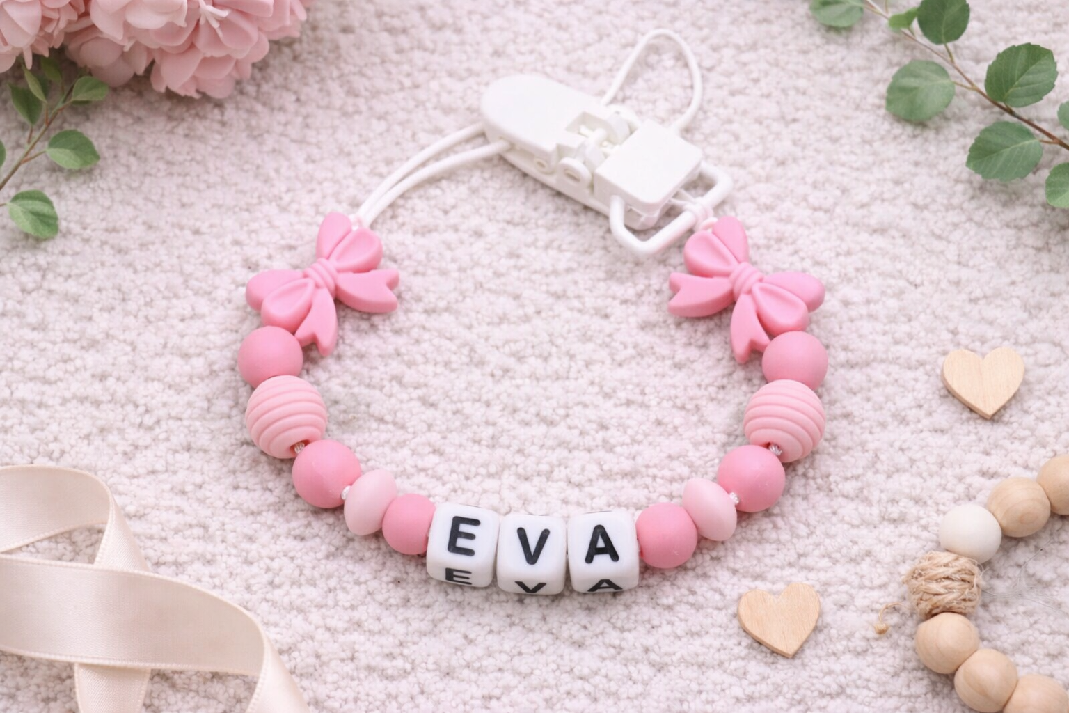 Personalized baby pacifier clip with pink silicone bows and beads spelling the name Eva on a soft cream textured background with floral accents.