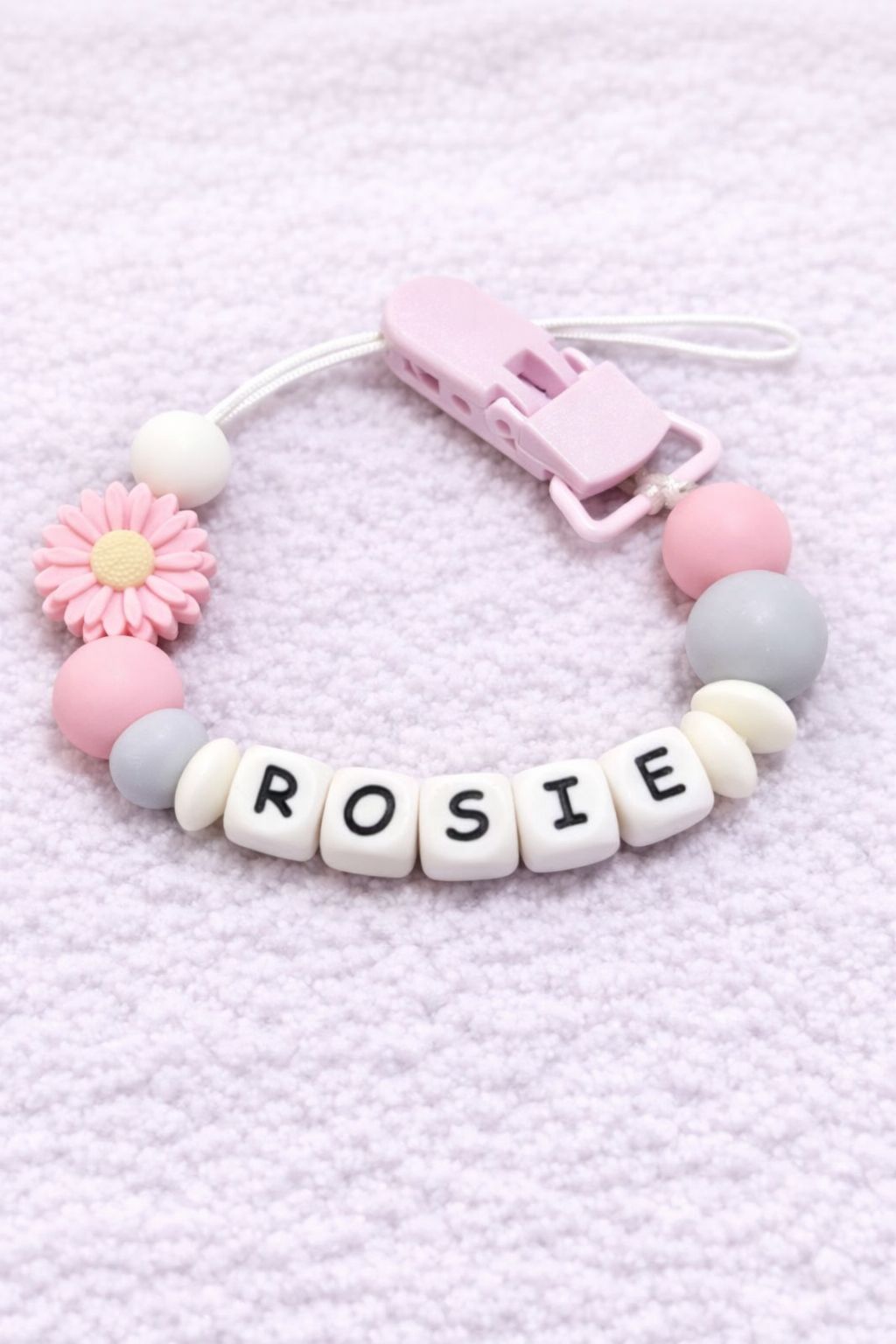 Baby dummy clip with 'ROSIE' beads on a light pink textured background