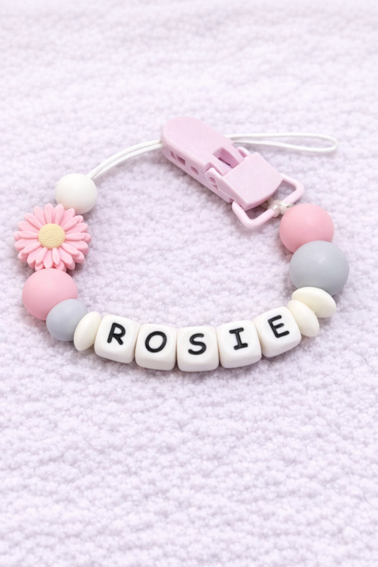 Baby dummy clip with 'ROSIE' beads on a light pink textured background