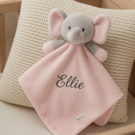 A personalized baby security blanket featuring a plush elephant head and a soft pink fabric body with the name 'Ellie' embroidered on it, resting on a textured cream pillow.