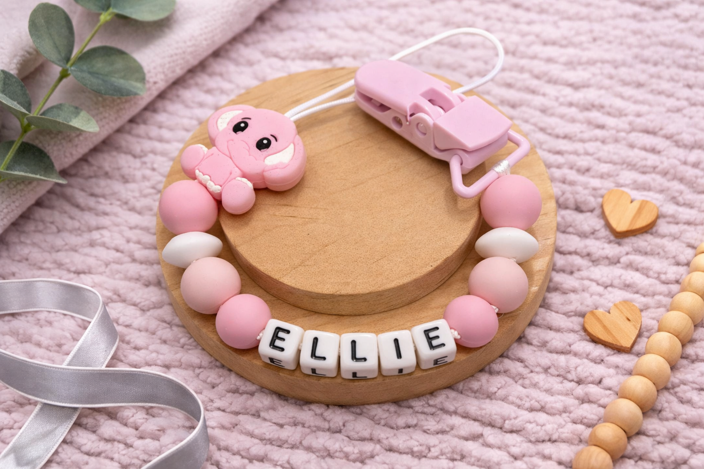 Pink elephant pscifier clip with 'ELLIE' beads on a wooden base, on a pink textured surface.