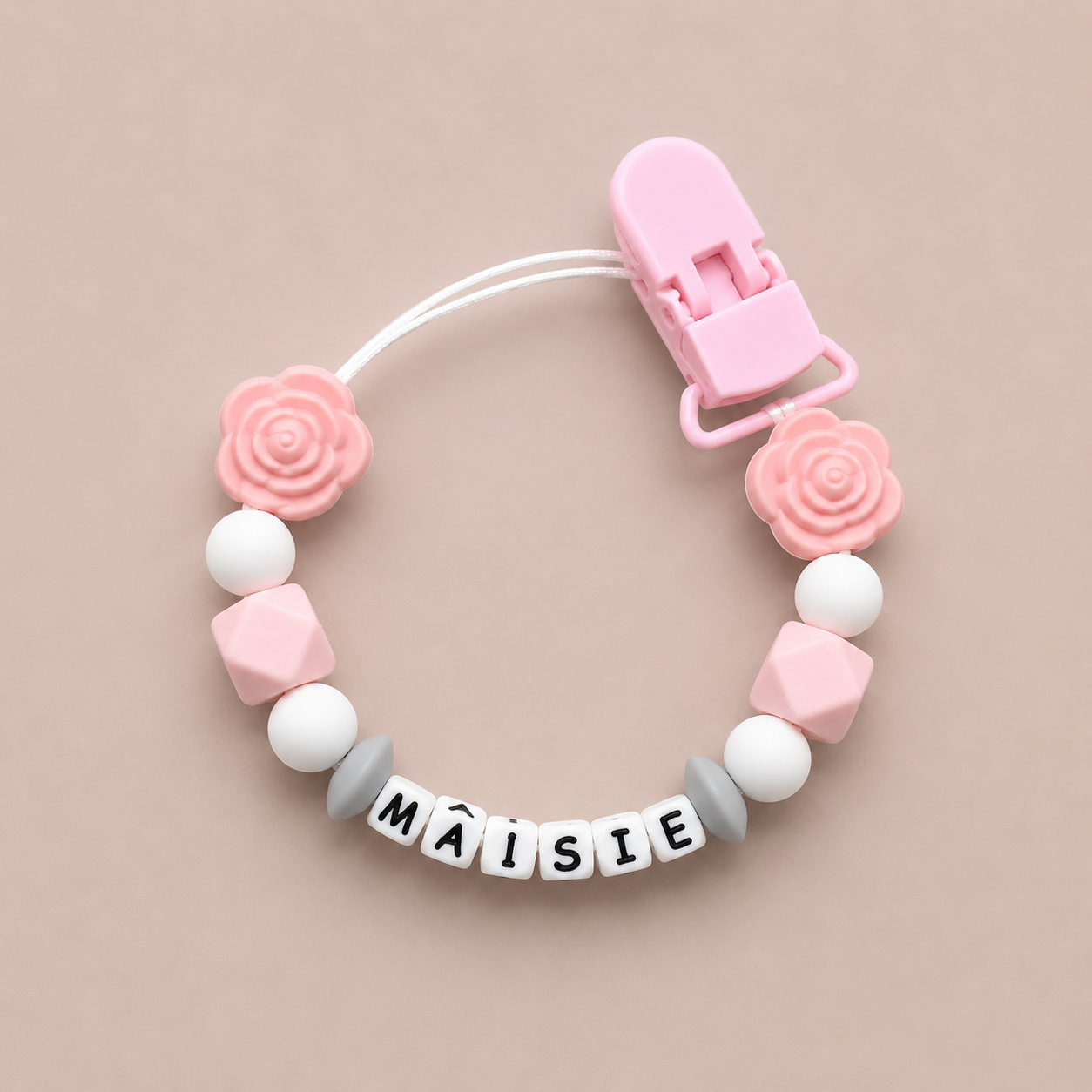 Pink and white pacifier clip with 'Máisie' on a beige background