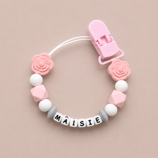 Pink and white pacifier clip with 'Máisie' on a beige background