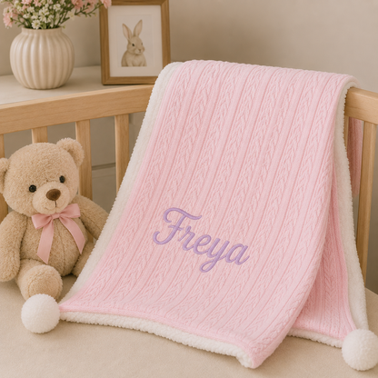 A pink cable-knit baby blanket with a white sherpa lining and the name 'Freya' embroidered in purple, draped over a wooden crib next to a teddy bear.