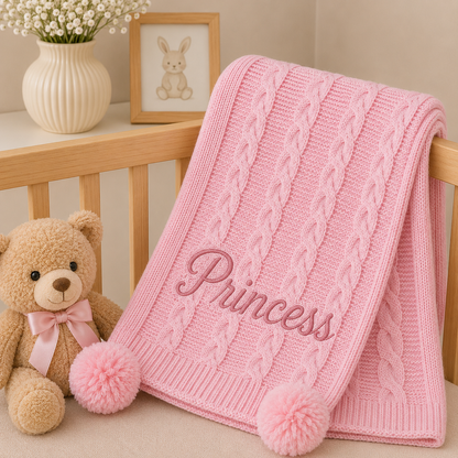 A pink cable-knit baby blanket draped over a wooden crib, featuring the name 'Princess' embroidered in pink script and two fluffy pom-poms at the corners. A teddy bear wearing a pink bow sits beside the crib, with a cream vase of flowers and a framed bunny picture in the background.