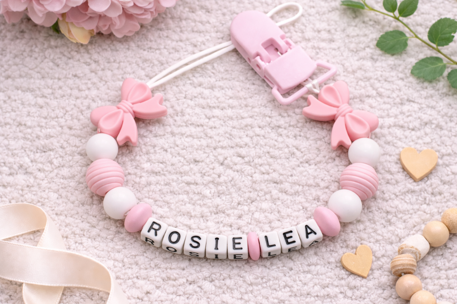 Personalised baby dummy clip with pink bows and white beads spelling “Princess” displayed on a wooden stand against a soft grey velvet background.