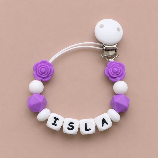 Baby dummy clip with purple and white beads spelling 'ISLA' on a beige background