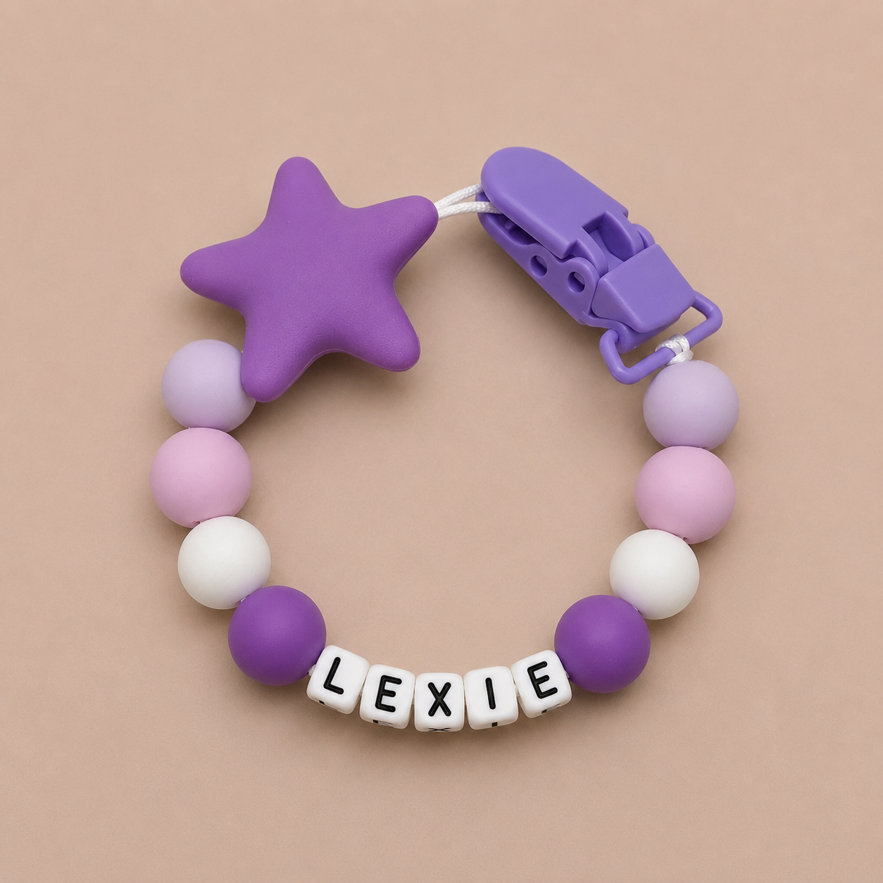 Purple star-shaped dummy clip with colorful beads and 'LEXIE' letter tiles on a beige background