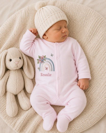 Newborn baby in a pink onesie with a rainbow design, lying on a textured surface next to a plush toy.