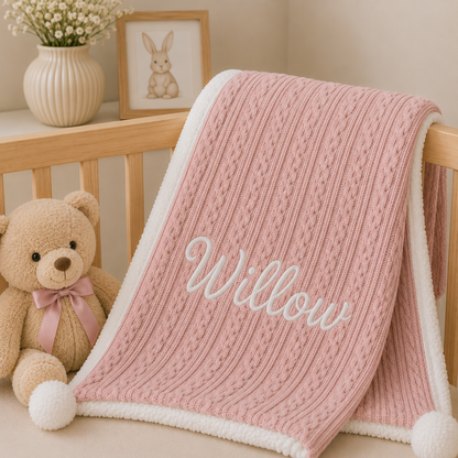 A pink cable-knit baby blanket with a white sherpa lining and the name 'Willow' embroidered in white script, draped over a wooden crib next to a teddy bear.