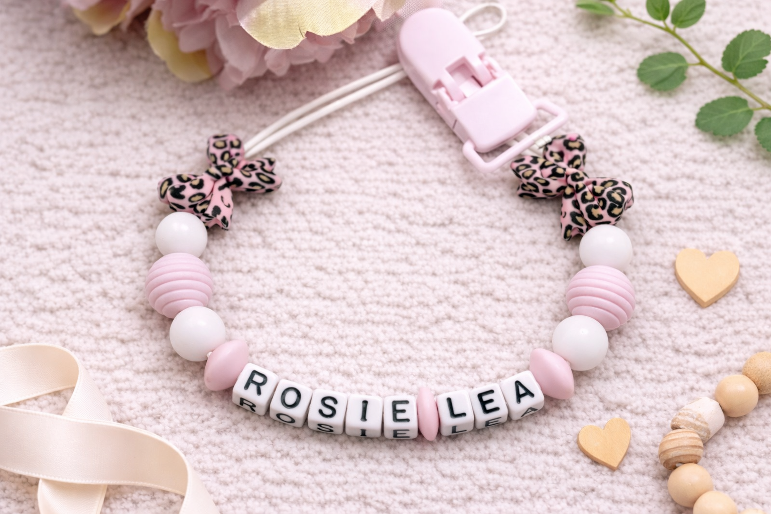 Flat lay image of a personalised baby pacifier clip featuring the name “ROSIE LEA” in white letter beads, surrounded by soft pink and white silicone beads and pink leopard print bow accents. The clip is styled on a textured cream background with blush flowers, wooden heart shapes, and greenery for a soft, feminine nursery-inspired look.