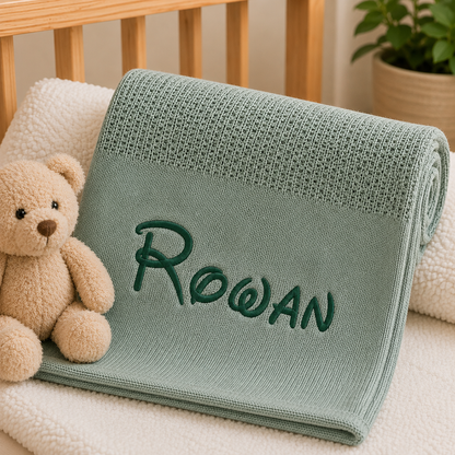 A personalized baby blanket in a dusty sage green color, rolled up and folded to show the name 'Rowan' embroidered in dark green script. The blanket is styled on a white fleece blanket next to a plush teddy bear, with a wooden crib and a potted plant in the background.