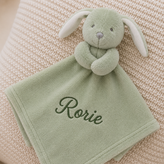 A personalized baby security blanket featuring a plush sage green bunny head attached to a matching soft square blanket. The blanket is embroidered with the name 'Rorie' in dark green script and is displayed on a textured cream knit blanket background.