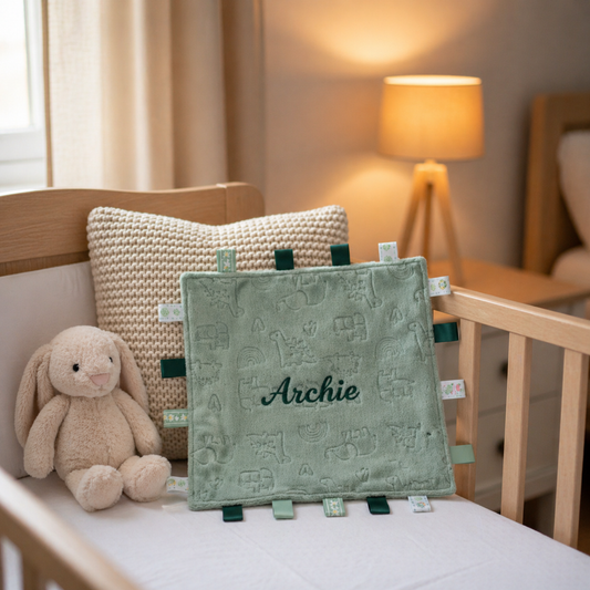 Green baby comfort blanket with 'Archie' embroidered, placed on a crib with a plush bunny toy and textured pillow.