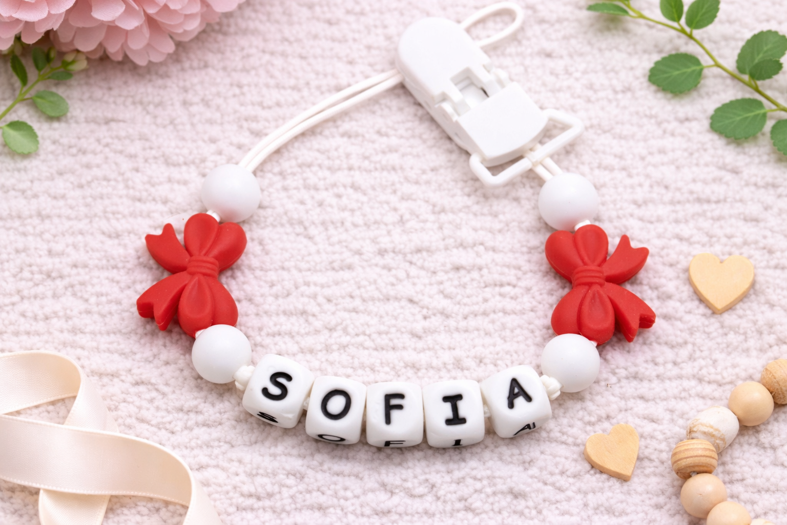 Flat lay of a personalised baby pacifier clip with the name “Sofia” in white letter beads, red bow silicone beads, and white beads on a soft cream textured background with floral accents.
