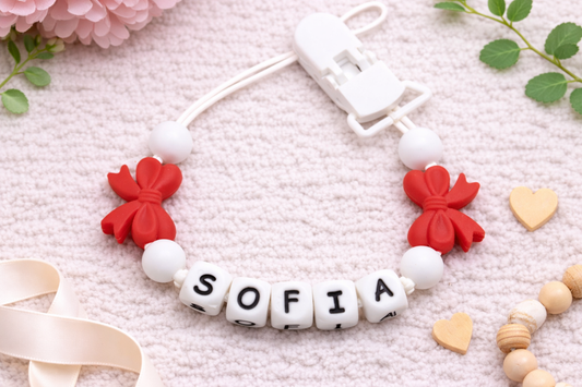 Flat lay of a personalised baby pacifier clip with the name “Sofia” in white letter beads, red bow silicone beads, and white beads on a soft cream textured background with floral accents.