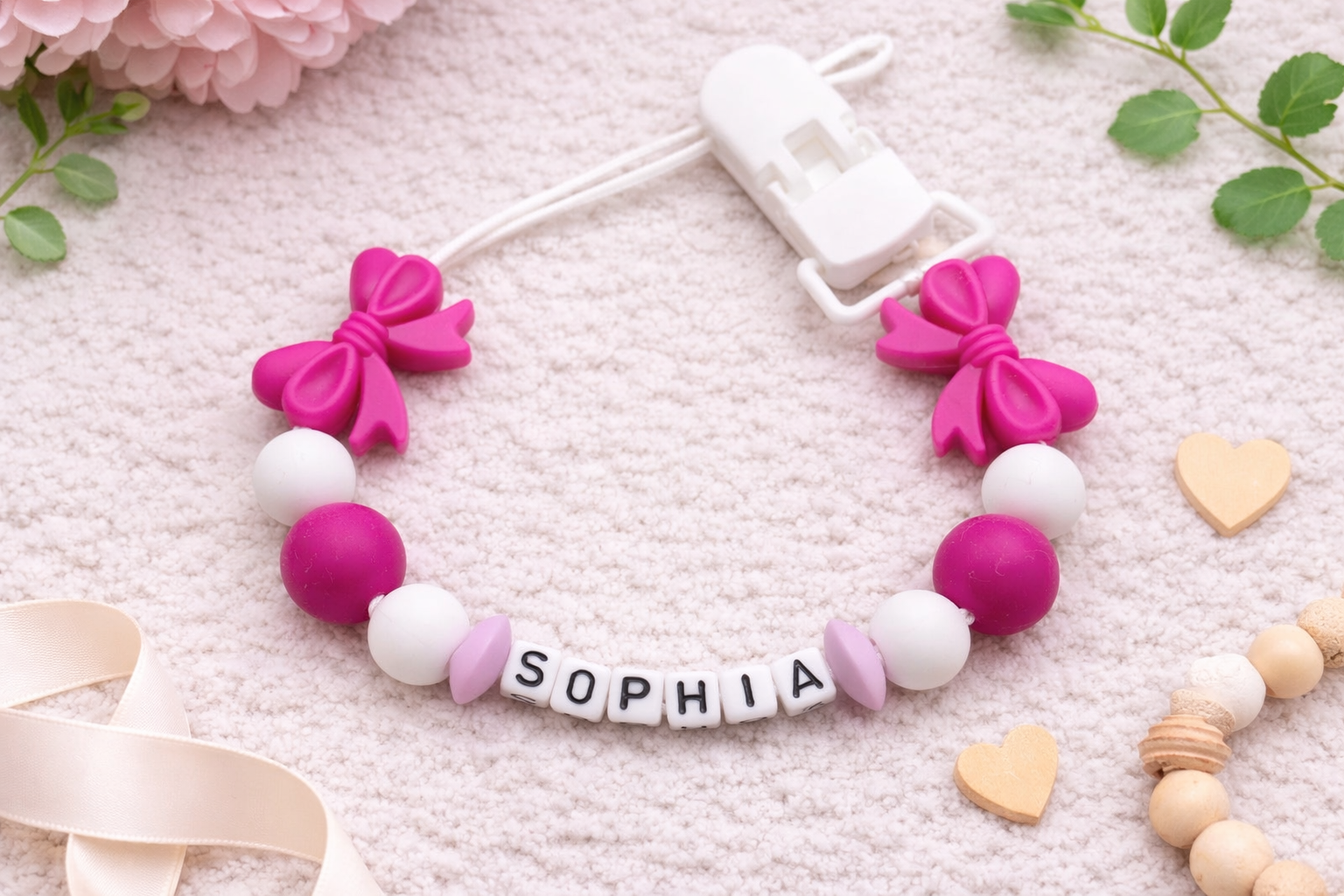 Personalised “Sophia” baby pacifier clip with bright pink bows and beads displayed on a soft cream flat lay background with flowers and wooden heart details.