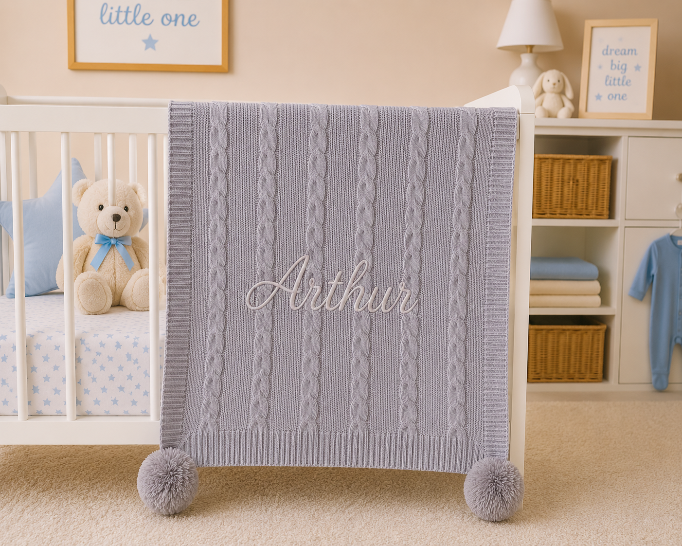 Baby crib with a gray cable knit blanket labeled 'Arthur', teddy bear, and baby clothes in a nursery.