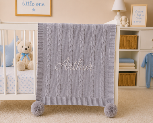 Baby crib with a gray cable knit blanket labeled 'Arthur', teddy bear, and baby clothes in a nursery.