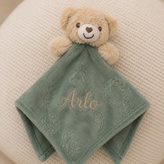 Teddy bear holding a green blanket with 'Arlo' embroidered on it, placed on a textured surface.