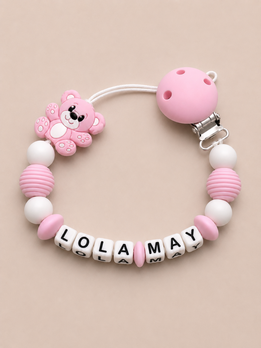Pink and white baby dummy clip with 'LOLA MAY' beads on a beige background