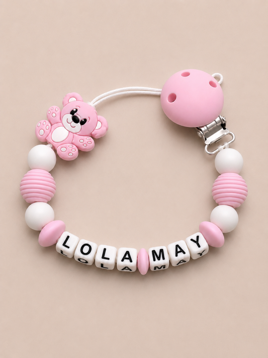 Pink and white baby dummy clip with 'LOLA MAY' beads on a beige background