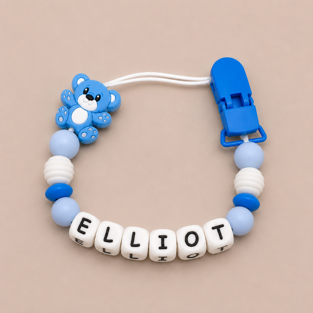 Blue and white pacifier clip with teddy bear charm and 'ELLiot' letter beads on a beige background