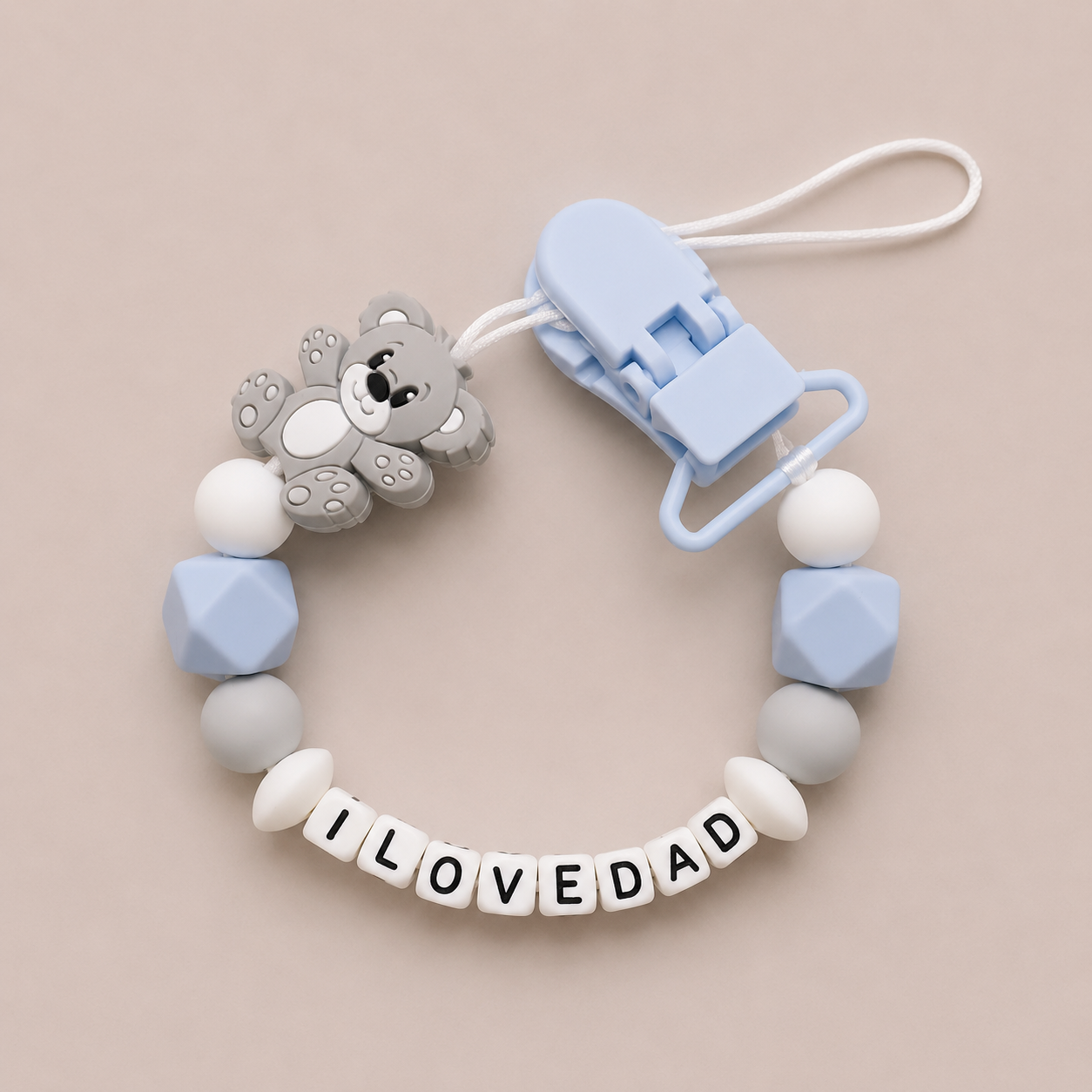 Baby dummy clip with teddy bear charm and 'ILOVEDAD' letters on a beige background