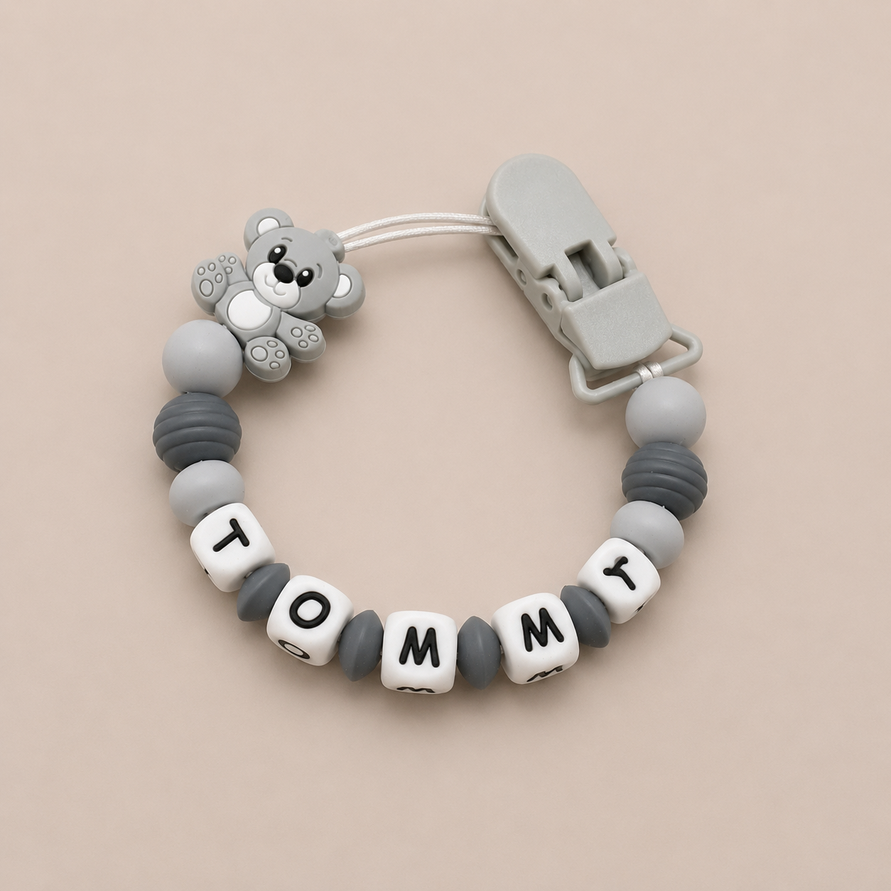 Baby dummy clip with teddy bear design and 'Tommy' beads on a beige background