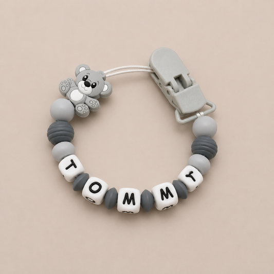 Baby dummy clip with teddy bear design and 'Tommy' beads on a beige background
