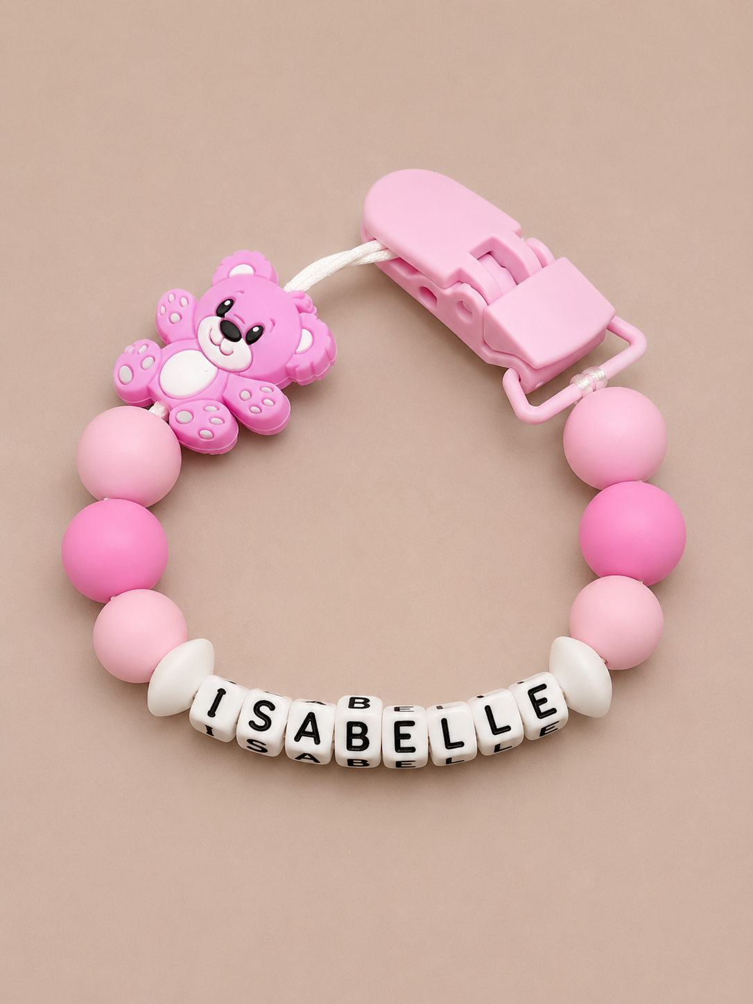 Pink pacifier clip with teddy bear design and 'ISABELLE' on beads against a beige background