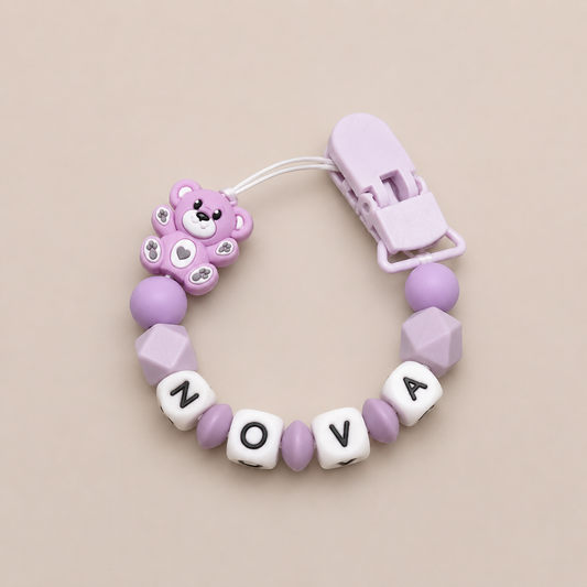 Purple pacifier clip with teddy bear charm and letter beads on a beige background