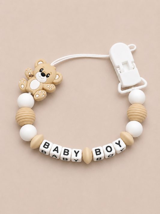 Baby dummy clip with teddy bear design and 'BABY BOY' beads on a beige background