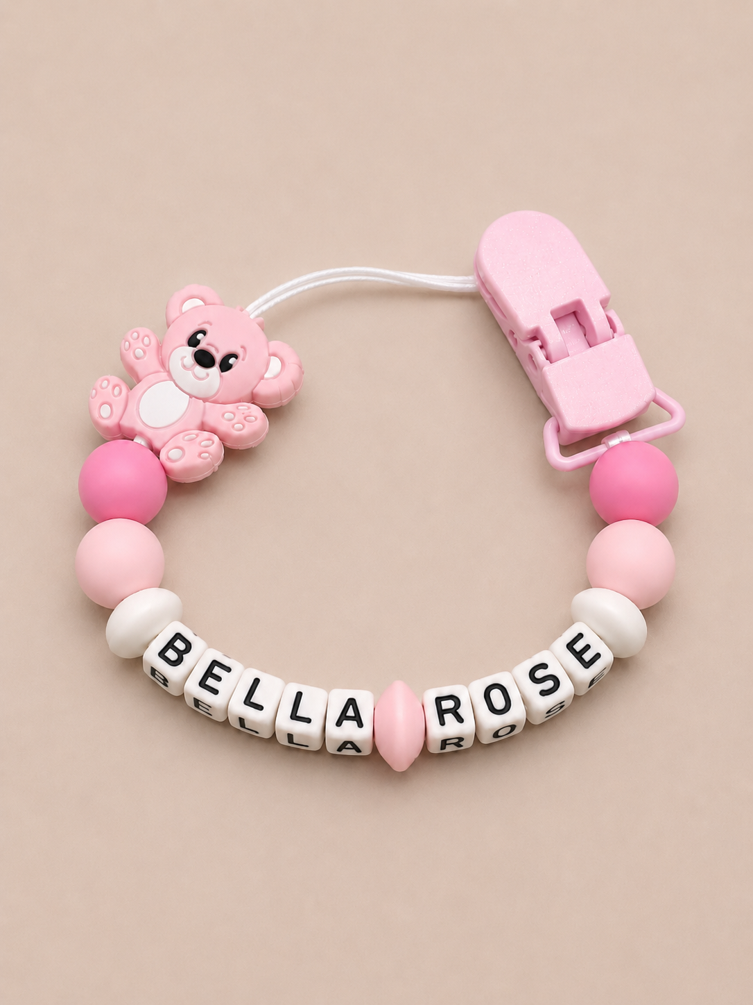 Pink pacifier clip with teddy bear charm and 'Bella Rose' letter beads on a beige background
