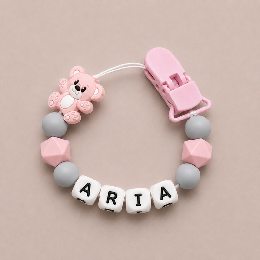 Baby dummy clip with pink bear charm and 'ARIA' letter beads on a beige background