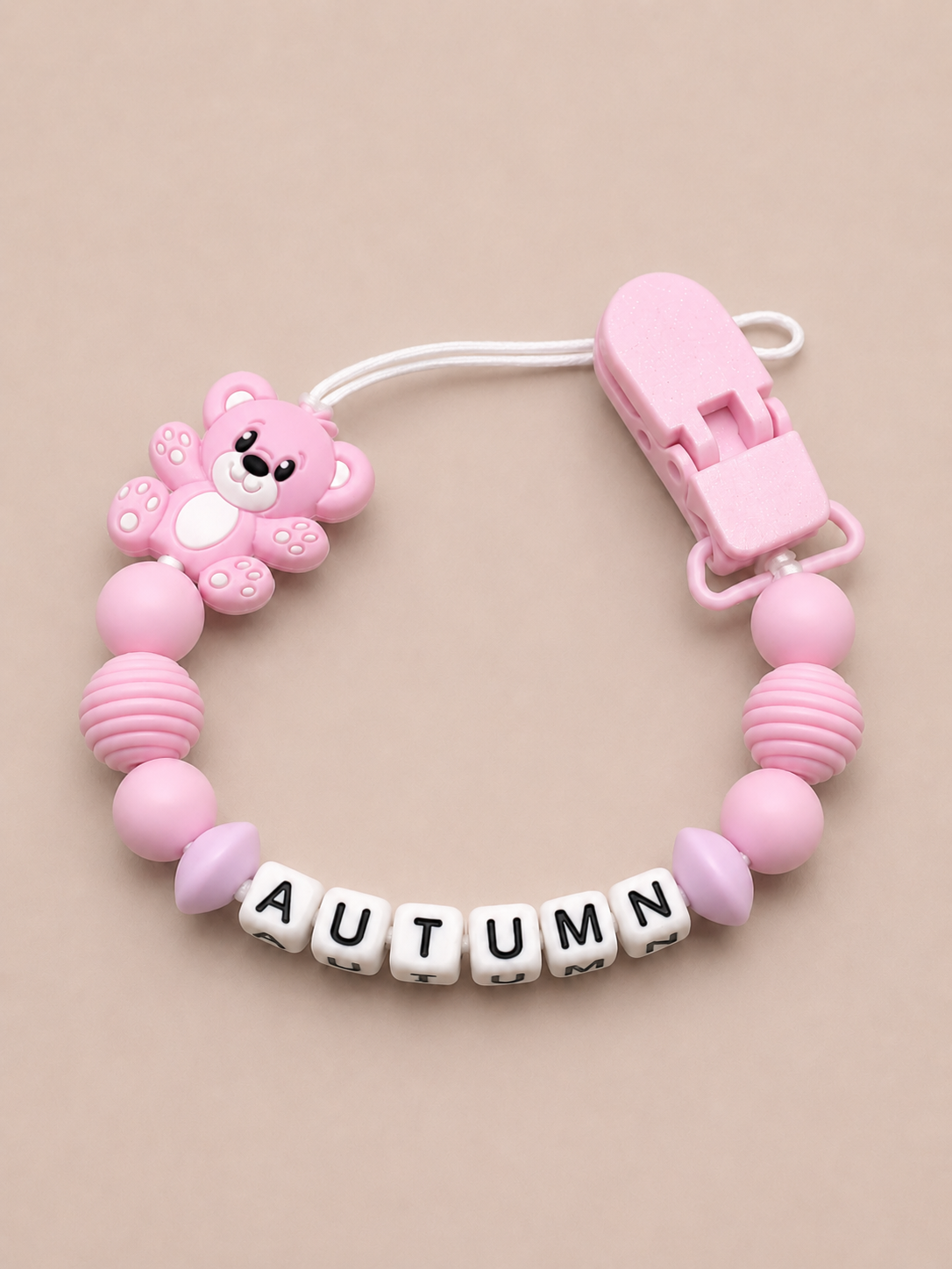 Pink baby dummy clip with a teddy bear design and 'AUTUMN' letters on a beige background