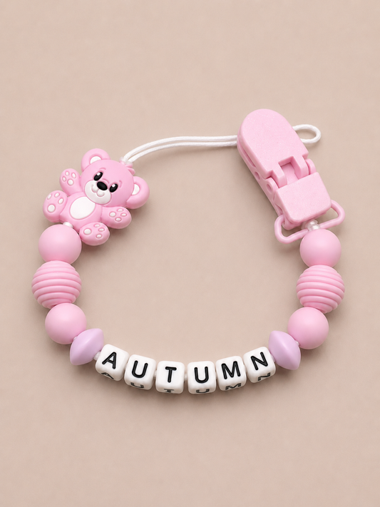 Pink baby dummy clip with a teddy bear design and 'AUTUMN' letters on a beige background