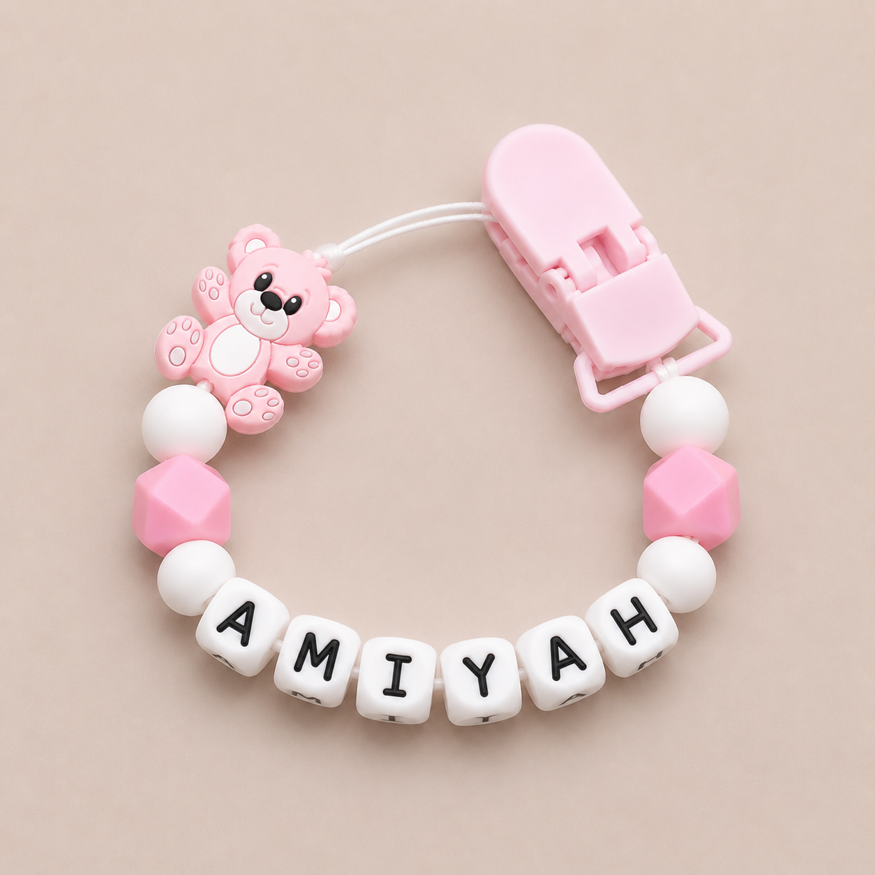 Pink and white baby dummy clip with a teddy bear charm and 'AMIYAH' letter beads on a beige background