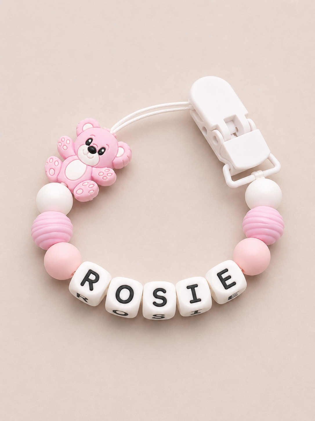 Pink and white dummy clip with letter beads spelling 'ROSIE' on a beige background