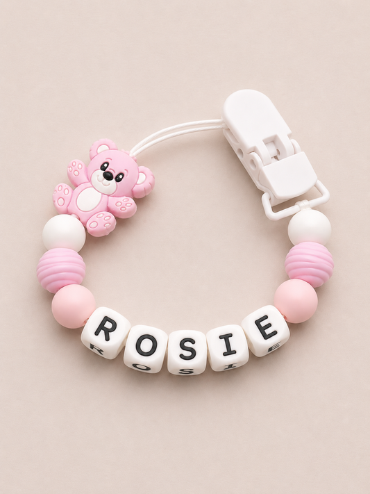 Pink and white dummy clip with letter beads spelling 'ROSIE' on a beige background