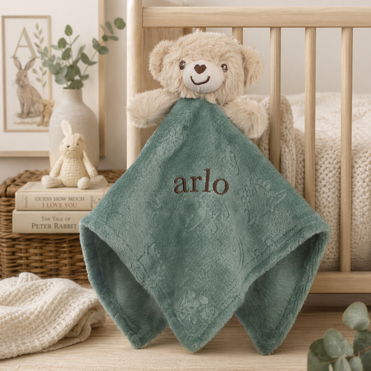 Green baby blanket with 'arlo' embroidered on it, draped over a teddy bear in a crib.