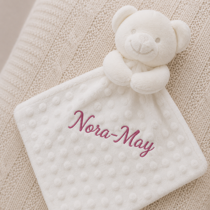 White teddy bear security blanket with 'Nora-May' embroidered on a beige background
