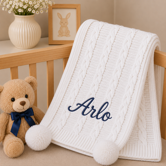 A white cable-knit baby blanket with two fluffy pom-poms and the name 'Arlo' embroidered in navy script, draped over a wooden crib next to a teddy bear and a small bunny picture frame.