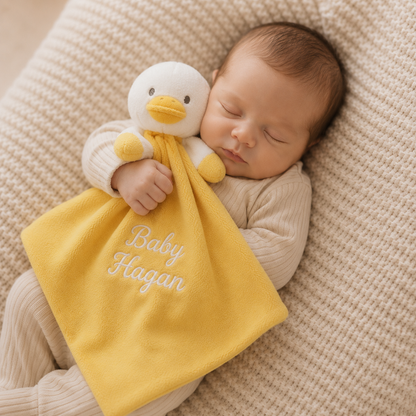 A sleeping newborn baby wearing a cream ribbed onesie, cuddling a yellow duck-shaped comforter blanket with the name 'Baby Hagan' embroidered on it, lying on a textured beige blanket.