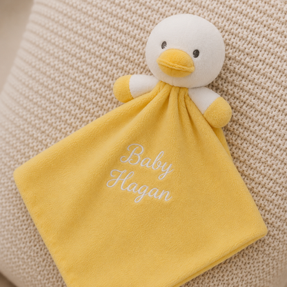 A yellow and white plush duck comforter with a square matching blanket attached, featuring the embroidered text 'Baby Hagan' in white script. The comforter is displayed on a textured beige woven blanket background.