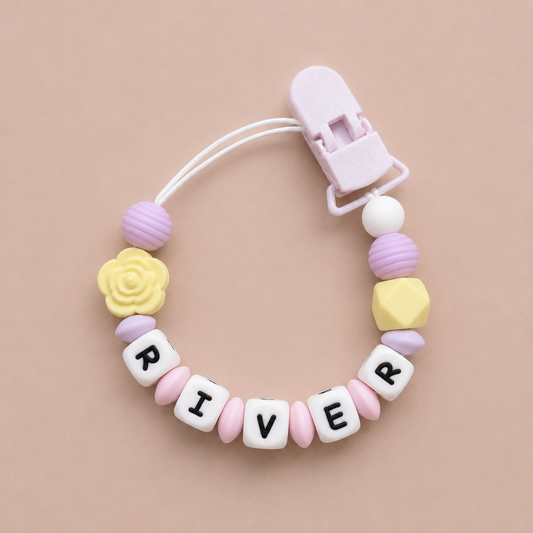 Pastel-colored pacifier clip with letter beads spelling 'RIVER' on a beige background