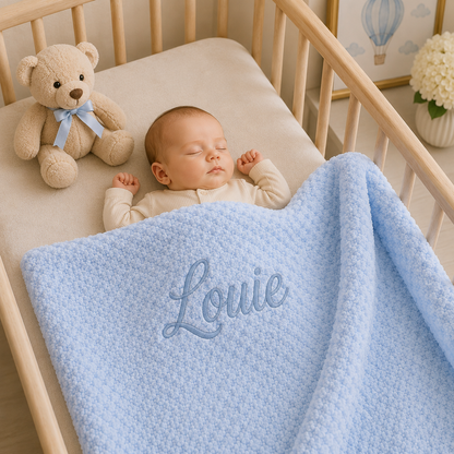A sleeping baby in a wooden crib under a light blue textured blanket with the name 'Louie' embroidered in blue script. A beige teddy bear with a blue ribbon sits on the crib mattress next to the baby.