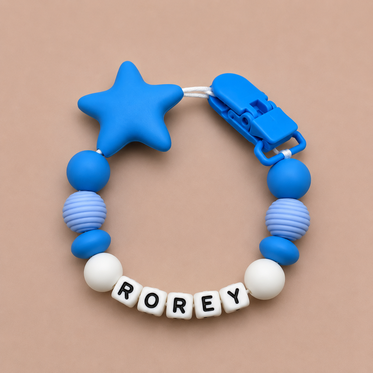 Blue dummy clip with star-shaped chewable and letter beads spelling 'ROREY' on a beige background